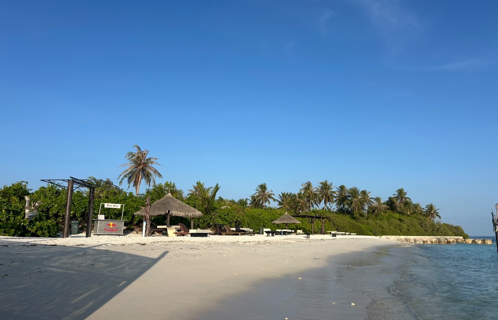 Kaashidhoo beach at the point: soft white sand, thatched sun shelters and palms beside a calm turquoise lagoon under a bright blue sky